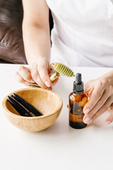 Brown plastic bottle for liquid cosmetics in woman's hands, green jade massage roller and wooden bowl with stone sticks on white table. Cosmetologist holding spin care products. 