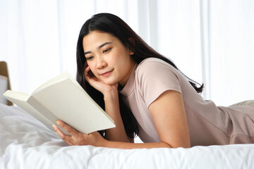 Obraz na płótnie Canvas Young Asian brunette woman lying down on the bed while reading a book in cozy and relax feeling