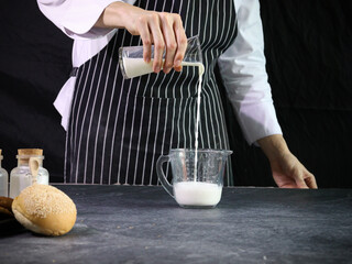 Bakery man pouring milk into a measuring jug in the kitchen at home, preparing bread dough, food...