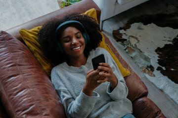 Mixed race female lying on sofa smiling while receiving exciting text on smartphone relaxing in modern lounge.
