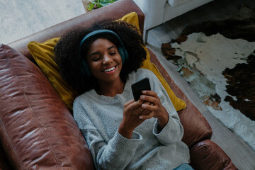 Mixed race female lying on sofa smiling while receiving exciting text on smartphone relaxing in...