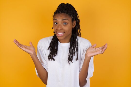 Clueless Young dark skinned woman with braids hair wearing white t-shirt over yellow background shrugs shoulders with hesitation, faces doubtful situation, spreads palms, Hard decision
