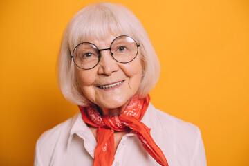 Closeup image of old cheerful woman with white hair and gray eyes wearing round glasses, white blouse, orange scarf or cravat. Woman standing isolated over orange background and smiling.