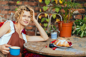 Blond haired middle age tired gardener in brown overalls relaxing after work at garden or backyard,...