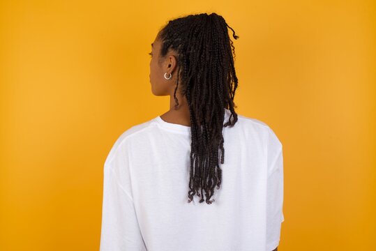 The back side view of a Young dark skinned woman with braids hair wearing casual clothes trendy isolated over yellow background Studio Shoot.