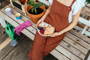 Blond haired middle age tired gardener in brown overalls relaxing after work at garden, holding plate with croissant over old brick wall sits at old bench in open air. Leisure, gardening concept.