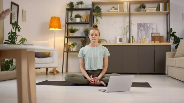 Tracking shot of young woman sitting in lotus pose on yoga mat and meditating while practicing with online instructor or teaching class via video call