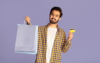 Smiling Indian man holding shopping bags and credit card on violet background