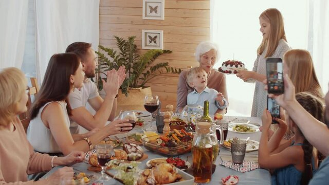 Cute little boy sitting on knees of grandmother while his sister bringing birthday cake on dinner table; kid blowing the candle while family clapping hands and taking pictures with smartphones