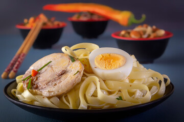 Noodles with pieces of meat and eggs in a black plate and dim sum with various traditional mushroom, soy and vegetable snacks with chopsticks on a blue table. Asian food concept