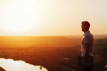 Young handsome man in headphones standing on top of the hill at orange sunset, listening to music. He holding a laptop. Copy space.