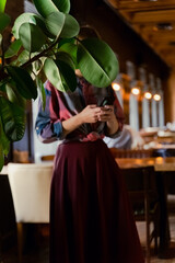 Faceless portrait of a young woman standing in restaurant hall holding smartphone, face hidden by leaves of a green plant