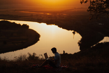 Silhouette of man working on laptop, he sitting on the peak of a mountain at sunset with river on background. Technology, internet, staying online in nature.