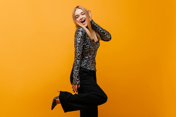 Ecstatic positive lady in high heel shoes standing on one leg. Cheerful girl in black pants dancing on yellow background.