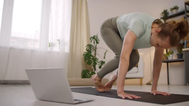Medium shot of young female yoga instructor doing downward facing dog pose on yoga mat while teaching online class during pandemic
