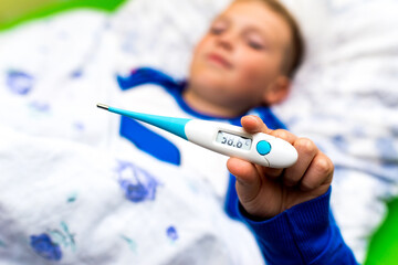 Close up ill young child or schoolboy, lying in bed shows a electronic thermometer, height of his fever