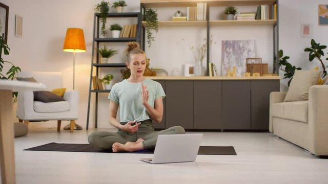 Tracking of young female yoga instructor sitting on mat at home and looking at laptop while talking and explaining poses during online class
