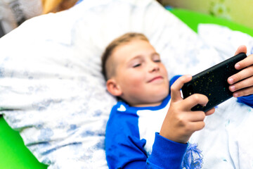 The sick child laying in bed looks into the smartphone and plays game while sick
