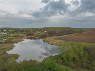 Fototapeta na wymiar Aerial perspective view of the pond surrounded by settlements, fields and forests under cloudy sky. Drone air view of the agricultural landscape with lake, village, trees and meadows. Summer day.