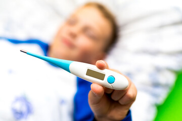 ill young child or schoolboy, lying in bed shows blank display of electronic or digital thermometer.Blurred background