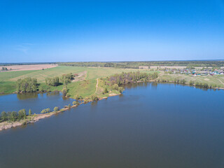 Aerial perspective view blue lake with shore covered by green grass and trees. Drone air view of the beautiful rural landscape with village on background under clear blue sky. Summer sunny day.