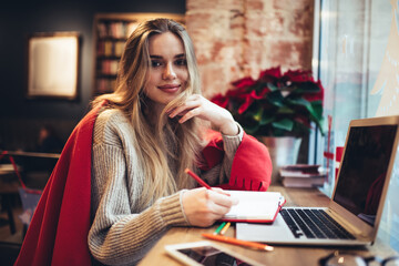 Smiling female freelancer taking notes in organizer and using computer in creative cafe