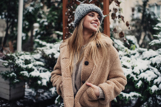 Cheerful Caucasian hipster girl 20 years old smiling at snowy street enjoying leisure time for winter walking, happy female tourist in eco coat and knitted hat enjoying cold season for travelling