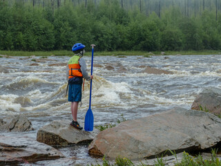 a boy with a paddle in a helmet with a life jacket standing on a large rock near a mountain river