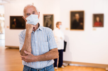 mature man in mask protecting against covid examines paintings on display in hall of art museum