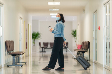 Fototapeta na wymiar Traveling young asian woman wearing face mask to protect covid19 virus and holding cup of coffee in her hand walking with suitcase bag at airport.