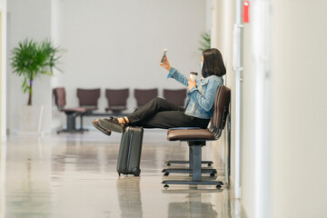 Fototapeta na wymiar Young asian businesswoman is photographing herself on cell telephone while is relaxing with take away coffee in the departure hall at airport. Copy space