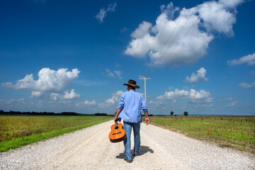 Man walking down one lane country dirt road wearing cowboy hat, carrying acoustic guitar, blue sky,...