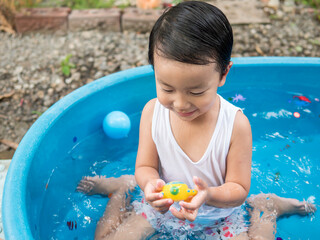 Asian cute child boy smiling while playing water in blue bowl with relaxing face and wet hair in rural nature. Young kid having happy moment in summer. Family activity at home and preschool concept.