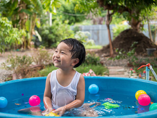 Fototapeta na wymiar Asian cute child boy smiling while playing water in blue bowl with relaxing face and wet hair in rural nature. Young kid having happy moment in summer. Family activity at home and preschool concept.