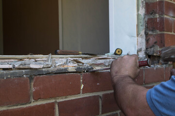 Workman using a hammer and chisel to remove old mortar from a window opening in a brick wall during...
