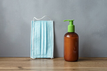 Flüssige Seife in einer brauen Flasche und ein blauer Mundschutz auf einem Holz Tisch. Graue Wand, Hygiene, Corona Virus.