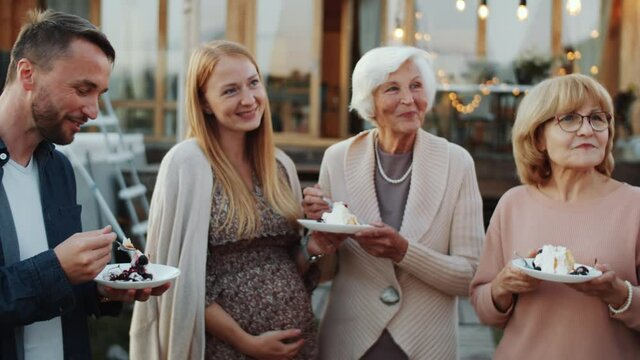 Cheerful mixed-age family members eating birthday cake, smiling and chatting while having dinner party outdoors in backyard decorated with lights