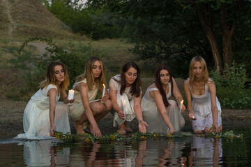 a group of young tangled girls in white dresses, launching flower wreaths in the water hold candles. On a natural background. A rustic style, a national holiday for young brides