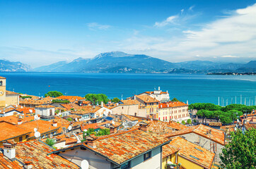 Naklejka na ściany i meble Aerial panoramic view of Desenzano del Garda town with red tiled roof buildings, Garda Lake water surface, Monte Baldo mountain range, Sirmione peninsula, blue sky background, Lombardy, Northern Italy