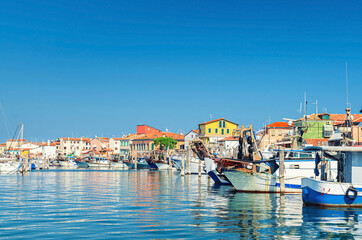 Fototapeta na wymiar Fishing ships and boats moored in port lagoon near seaside of Sottomarina town with row of colorful buildings in summer day, blue sky background, Veneto Region, Northern Italy