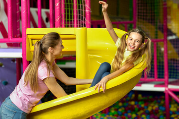 Fototapeta na wymiar Young mom with her daughter having fun on playground slide at indoor kids center