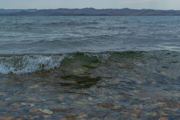 bright emerald green waves of lake baikal in the light of the sun in summer, blue mountains on the horizon, pebbles in water, coast
