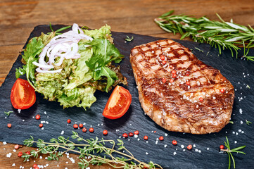 Grilled beef steak with fresh green salad leaves, sliced tomatoes and onions, red pepper and salt on stone tray