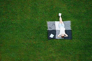 Young attractive caucasian woman with closed eyes lying on the blanket on green grass and listen to...