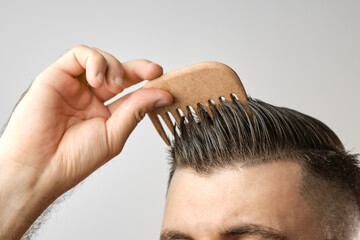 Close up man brushing his hair with wooden comb on the grey background. Treatment against hair lost and dandruff concept. Cosmetic products for men. Barbershop advertising.  - obrazy, fototapety, plakaty