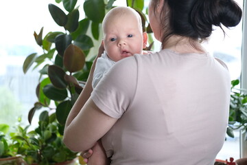 A beautiful young mother holds her newborn son in her arms by the window. Close-up portrait of a...