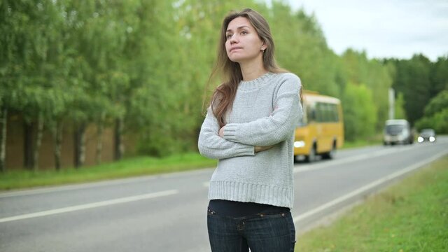 The camera zooms in on a woman who is waiting for a ride or a bus on the road outside the city