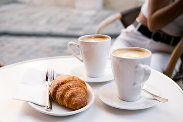 Two lattes coffee in white cups on a saucer and a croissant in a white plate, a fork and a knife on a summer terrace in a cafe. Girl with a phone in the background.