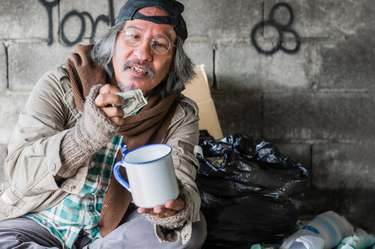 Happy homeless senior man from banknote and money by donation. Homeless old man wear sweater and blanket sitting on cardboard seeking help and food beggar because hungry from people on walkway street.