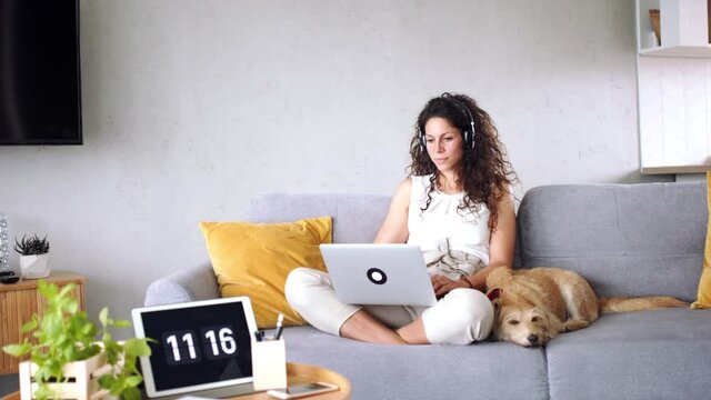 Woman with pet dog sitting indoors at home, using headphones and laptop.